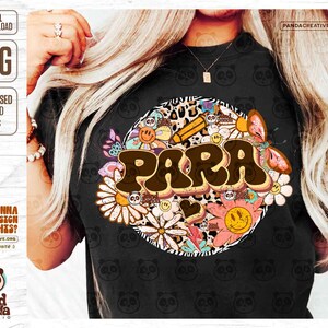 K&ouml;nnte beinhalten: Ein schwarzes T-Shirt mit einem farbenfrohen Grafikdesign, das das Wort "PARA" in einer Retro-Schriftart zeigt, umgeben von Blumen, Schmetterlingen und anderen skurrilen Elementen. Das Design ist auf einem Leopardenmuster-Hintergrund platziert.