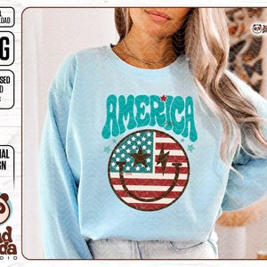 May include: A light blue sweatshirt with a graphic design of a smiley face with the American flag pattern. The text "AMERICA" is in a retro font above the smiley face.