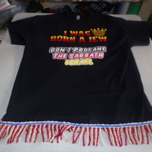 Pode incluir: Camiseta preta com o texto "I was born a Jew Don't profane the Sabbath Israel" em letras amarelas, vermelhas e brancas. A camiseta tem uma bainha de franjas douradas, vermelhas e azuis.