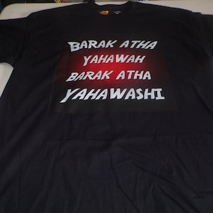 May include: Black t-shirt with white text that reads "BARAK ATHA YAHWAH BARAK ATHA YAHAWASHI" in a rectangular design with a red gradient background. The shirt is made of a soft material.