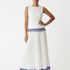 Puede incluir: Un vestido largo plisado blanco sin mangas con flecos azules y morados en la cintura y el dobladillo. El vestido se combina con tacones blancos. La modelo está de pie con una mano en la cadera.