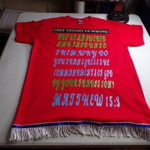 Può includere: Maglietta rossa con bordino di frange bianche e nastro blu. La maglietta ha una stampa di testo verde, blu e viola che dice "THEY TAUGHT US WRONG BUT HE ANSWERED AND SAID UNTO THEM WHY DO YOU TRANSGRESS THE COMMANDMENTS OF GOD BY YOUR TRADITION? MATTHEW 15:3"