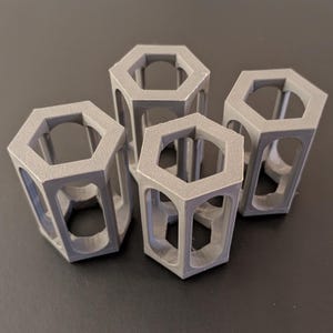Puede incluir: Cuatro objetos grises en forma hexagonal con lados y partes superiores abiertas. Los objetos tienen una superficie texturizada y están dispuestos sobre una superficie oscura. El diseño presenta una estructura similar a un panal, lo que sugiere un tema geométrico o industrial.