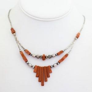 May include: A two-strand necklace with silver-tone metal and reddish-brown rectangular and oval beads. The necklace has a fan-shaped design at the bottom and is displayed on a white mannequin.