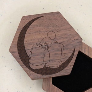 May include: Hexagonal wooden ring box with a hinged lid. The lid features an engraved design of a couple seated on a crescent moon. The interior of the box is lined with black fabric. A small, round, gold-colored clasp secures the lid.