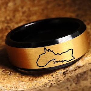 Puede incluir: Un anillo negro y dorado con un contorno de la isla de Guam y la palabra "Guam" inscrita en la banda.