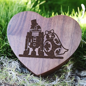 May include: A heart-shaped wooden ring box with a laser-engraved design of R2-D2 and BB-8 dressed as a wedding couple. The box is made of walnut wood and has a natural finish.