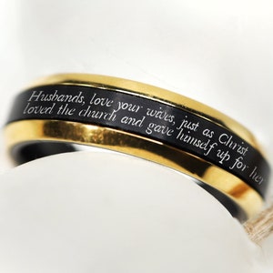 May include: A close-up of a gold and black ring. The ring has a gold band with a black center band. The text "Husbands, love your wives, just as Christ loved the church and gave himself up for her" is inscribed on the black band.