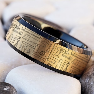Puede incluir: Un anillo negro y dorado con un diseño de varias herramientas en la banda.