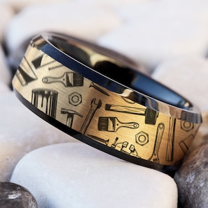 Puede incluir: Un anillo de carburo de tungsteno negro y dorado con un borde biselado y un diseño de varias herramientas en la banda.
