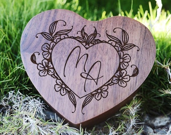 May include: A heart-shaped wooden ring box with a floral design and the initials "M & A" engraved inside. The box is sitting on a bed of green grass.
