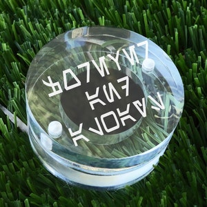 May include: A clear plastic disc with white lettering that reads "KJO KIV KIV7 Y7M7" on a black background.