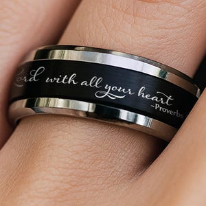 以下が含まれることがあります： 「Trust in the Lord with all your heart - Proverbs 3:5」と刻まれた黒とシルバーのリング。指にはめられています。