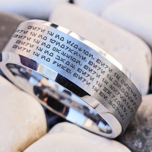 Puede incluir: Un anillo de plata con una banda ancha grabada con un patrón repetitivo de símbolos. El anillo está descansando sobre un lecho de piedras blancas.