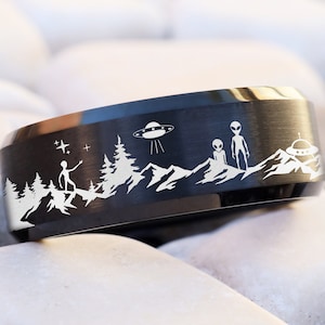 May include: A black tungsten carbide ring with an engraved design of a UFO, aliens, and a mountain landscape. The ring is a brushed finish.