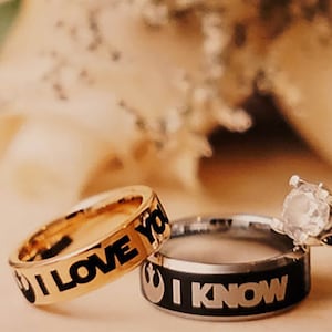 May include: Two gold wedding bands with black lettering. The band on the left says "I Love You" and the band on the right says "I Know". The bands are on a white background with a bouquet of white roses.