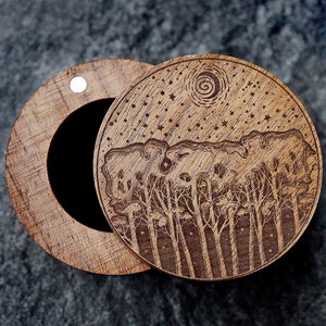 May include: A wooden box with a hinged lid. The lid is decorated with a laser-engraved design of a forest scene with trees, stars, and a crescent moon. The box is closed, but the lid is slightly ajar.