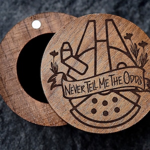 May include: A wooden ring box with a laser engraved design of the Millennium Falcon from Star Wars. The box has a small floral design and the text "Never Tell Me The Odds".
