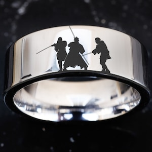 May include: A silver ring with a black silhouette of three figures wielding lightsabers. The figures are facing forward and appear to be in a battle stance.