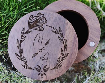 May include: A round wooden ring box with a floral design and the initials "M & A" engraved on the lid. The box is open and the inside is lined with black felt.