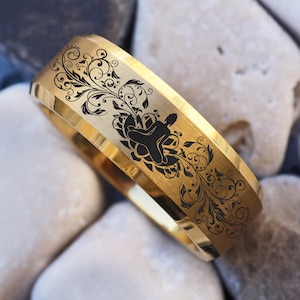 May include: A gold-colored ring with a black floral design and a geometric emblem. The ring is set against a background of white and gray stones, creating a contrast between the shiny metal and the natural textures.