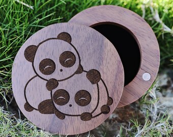 Puede incluir: Una caja de madera con tapa abatible, que presenta un diseño grabado con láser de dos pandas abrazándose. La caja es de madera marrón oscura y tiene un pequeño cierre plateado.