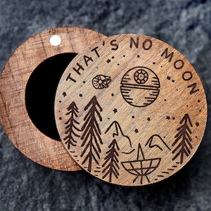 Pode incluir: Uma caixa de madeira com uma tampa que se abre para revelar um pequeno compartimento. A tampa é decorada com um design gravado a laser de uma cena de floresta com uma Estrela da Morte e o texto "That's no moon."