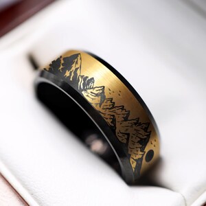 May include: A black and gold wedding band with a mountain range design. The band is in a ring box.