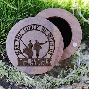 Pode incluir: Uma caixa de anel redonda de madeira com tampa. A tampa tem um desenho gravado de um casal correndo com o texto "Que a força esteja connosco" e "Mr & Mrs".