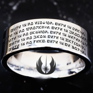 May include: A silver ring with a black inscription in Aurebesh, the language of the Star Wars universe. The inscription reads "The Force will be with you, always." The ring also features a silver Jedi symbol with a lightsaber and a star.