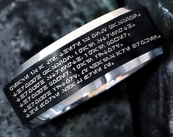 May include: A black and silver ring with a band of black metal engraved with a series of symbols in a language that resembles Aurebesh, the fictional alphabet used in the Star Wars universe. The inscription reads: "ルミフロロG N100 115 ミ・ルミフロロブ11000017. ルミプロロ M7,1 DK10 Y15ZV MKM 12 K J1V MN100. ルミプロコロミ ¥144077.41M X7M 370 ルミ 川ㄥ 7MM V".
