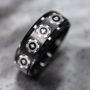 Puede incluir: Un anillo de carburo de tungsteno negro con una banda plateada que presenta cuatro diseños de fichas de póquer con diferentes palos de cartas.