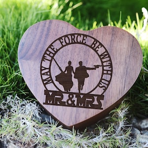 Pode incluir: Uma caixa de alianças de madeira em forma de coração com um design gravado a laser. O design apresenta um casal de mãos dadas, um sabre de luz e um blaster. O texto diz "Que a Força esteja connosco" e "Mr. & Mrs."