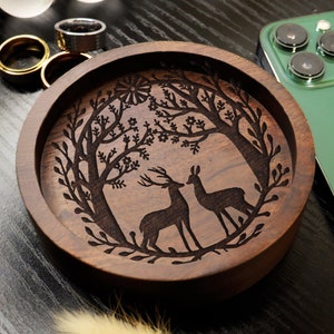 May include: A wooden trinket dish with a carved design of two deer standing in a forest. The dish is dark brown and has a smooth, polished finish.