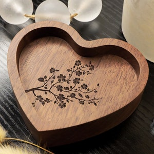 May include: A heart-shaped wooden ring dish with a cherry blossom tree design. The dish is made of dark wood and has a smooth finish.
