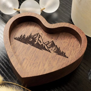 May include: A heart-shaped wooden ring dish with a laser-engraved design of mountains and trees. The dish is made of dark wood and has a smooth finish.