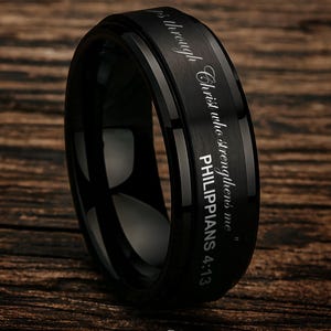 May include: A black ring with the inscription "I can do all things through Christ who strengthens me" and "Philippians 4:13". The ring has a polished finish and is set against a wooden background.