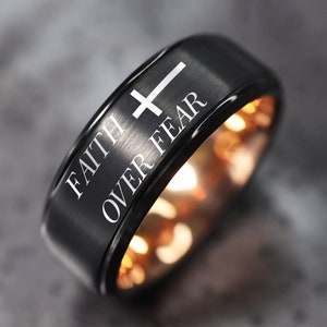 May include: A black tungsten carbide ring with a rose gold inner band. The ring has a brushed finish and features a white cross and the words "Faith Over Fear" engraved on the top.
