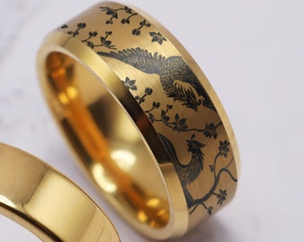 May include: A pair of gold wedding bands with a black etched design of birds and flowers. The band on the left is plain, while the band on the right has a detailed design.