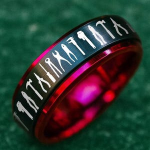 May include: A red and black ring with a silver design. The ring has a black band with a repeating pattern of silver silhouettes. The inner band of the ring is a vibrant red color. The ring is set against a green background.