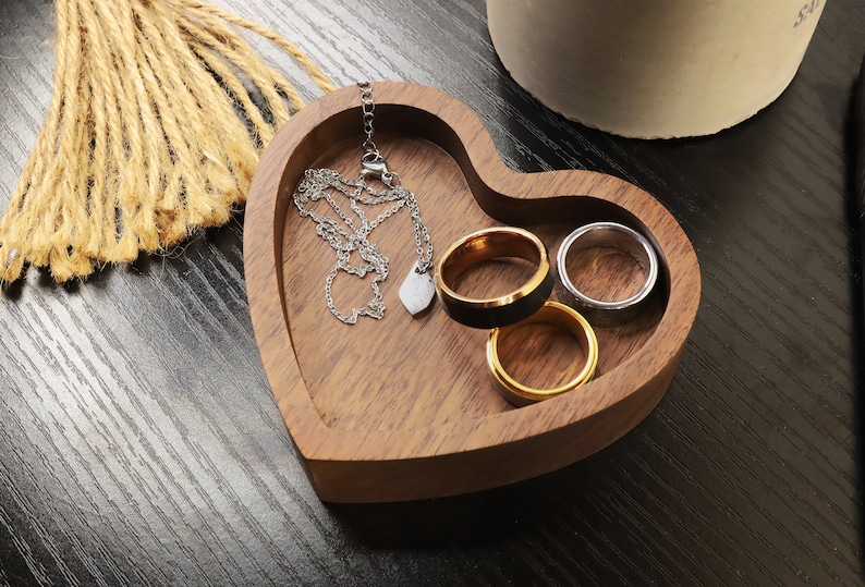 May include: A heart-shaped wooden trinket dish with a silver chain necklace and four rings. The rings are gold, silver, and black.
