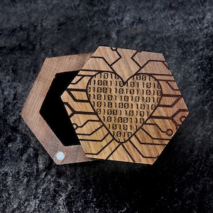May include: A hexagonal wooden ring box. The lid has a laser-engraved design of a heart made of binary code on a circuit board pattern.