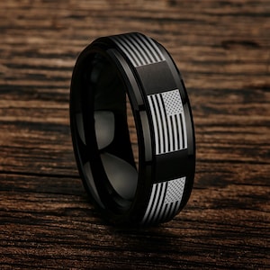 May include: A black ring with a polished finish. The ring features a design of the American flag in white and gray. The flag design is repeated around the ring's band. The ring is set against a wooden background.