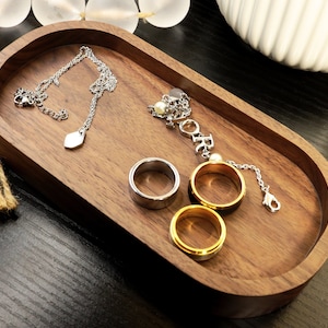 May include: A wooden oval tray with a silver necklace, a silver chain with a charm that says "FOREVER", and three rings. Two of the rings are silver and one is gold.