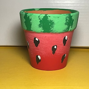 Può includere: Un piccolo vaso di terracotta dipinto per assomigliare a un'anguria. Il vaso è rosso con strisce verdi intorno alla parte superiore e semi neri dipinti sul davanti.