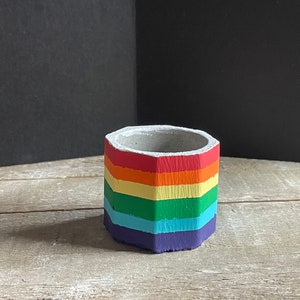 May include: A hexagonal concrete planter painted with rainbow stripes in red, orange, yellow, green, blue, and purple.