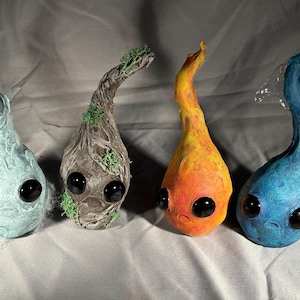 May include: Four small, hand-crafted figurines with googly eyes. Each figurine is shaped like a teardrop and has a different color and texture. The figurines are made to represent the four elements: water, earth, fire, and air.