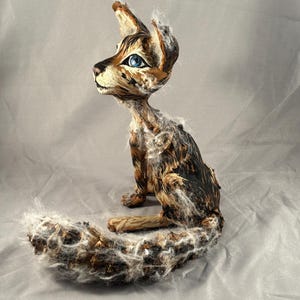 May include: A handcrafted sculpture of a cat with a slender body and large ears. The cat is primarily brown and black with white accents, and has striking blue eyes. The tail is long and fluffy, and the cat is posed in a seated position.