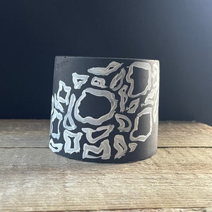 May include: A cylindrical black planter featuring a hand-painted white abstract pattern of irregular, thick-lined shapes. The vessel has a smooth, matte finish and a wide, open top, showcasing a stark contrast between the dark base and white paint.
