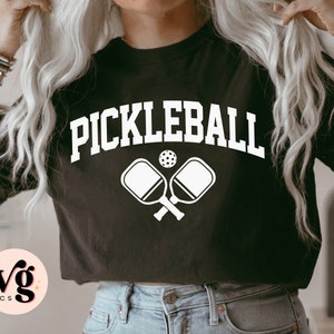 May include: Black t-shirt with the word "PICKLEBALL" in white, arched lettering. Below the word are two crossed pickleball paddles and a pickleball. The shirt has a crew neck and short sleeves. The logo "svg basics" is in the lower left corner.